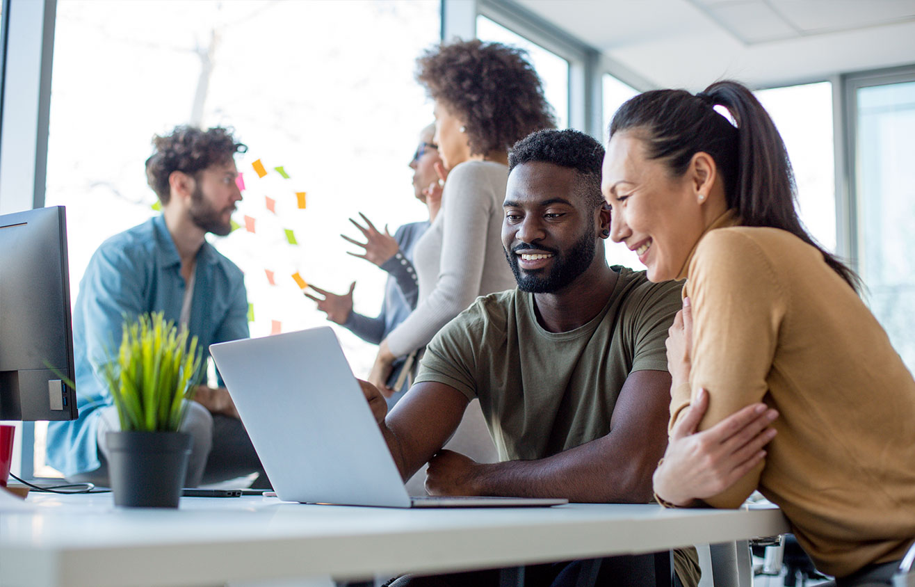 Man and woman at a desk in an office smiling while looking at a laptop